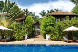 A tropical boutique hotel in Battambang, Cambodia, featuring a serene blue pool surrounded by lush greenery, palm trees, and traditional wooden villas with terracotta roofs, creating a tranquil and luxurious retreat.