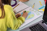 A child wearing a yellow sweater is drawing the planet Earth on a notebook. The word “Earth” is colored in green.