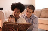 Interracial heterosexual couple of white male and brown-skinned female with short curly hair. The mom is holding a 2 year old on her lap and they’re reading her a story from a big, brown, old-looking book. They are sitting on a living room floor next to the couch.
