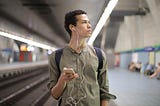 A young man walks through a transit station while listening to music through wired headphones.