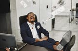 Man in a suit sitting at a desk with his feet up, smiling joyfully as hundred-dollar bills fall around him in an office setting.