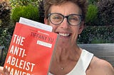 Photo of Karen Catlin smiling while holding a copy of Tiffany Yu’s new book, The Anti-Ableist Manifesto. It has an orange cover with white and gray text. Behind Karen is a perfectly blue sky and some rose bushes.