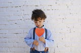 small boy holding a red, paper heart, standing in front of a white brick wall