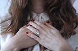 A brunette, white woman with her hands with red painted nails and a rings on her heart space