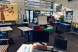 A desk with computer and laptop sits in the front of the picture. Behind, you can see a classroom with tables, chairs.