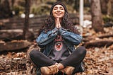 Smiling woman sitting in forest in jean jacket and hiking boots, hands in prayer posture under chin.