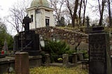 Foreground is individual graves with headstones. In background is a knoll with a small mausoleum with more individual headstones beyond it.