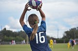 Photo of a girl throwing a soccer ball. It is shot from the back and other players are out of focus.