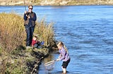 woman and two kids fishing. Adult has rod, one child sits next to her , other in stream with net