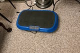 Blue LifePro vibration plate exercise machine with a black textured surface and control panel, placed on beige carpet near a lamp and ventilation grate.