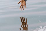 An arm extended from a boat, the hand reaching toward its reflection in the water.