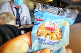 A photo from the first class seat of an airplane, author holding up a cookie in a package near with the Steward in the background. There’s an image of a little man on the cookie wrapper that looks uncannily like the Steward