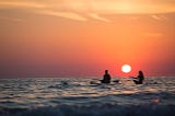 Two people sit on paddle boards in the ocean as the sun sets behind them