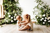 Little girl holds hands of baby while both sit on the floor with white roses in the background