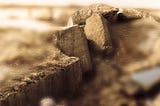 A heavily modified photograph showing the shattered remains of a bunker, consisting of huge concrete blocks, lying in a large crater. The image has been given an overall monochrome tint and many parts of it have been blurred to concentrate on the blocks.