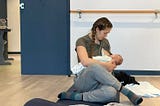 A woman on a yoga mat sits in cow face pose while nursing a baby