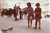 A sepia-toned photograph of a young woman and her son kneeling before some pigeons, preparing to feed them