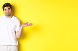 A clueless looking man in a white t shirt, shrugging and looking puzzled as if he has no idea what is going on. Bright yellow background.