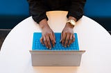 Uma mulher negra digita em um notebook de teclados azuis, vemos suas mãos, relógio dourado e blusa preta de mangas. Ela tem o notebook apoiado em uma mesa redonda branca.