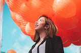 A woman in a business suit looks up at a net full of balloons above her. An excited/optimistic smile is on her face.