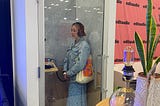A photo of a person with blonder wearing a jean jacket and long blue and white skirt, wearing headphones and speaking in the editaudio pop-up sound booth.