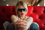 A slim, pretty girl with a blonde bob and scarlet lipstick sits on a red diner banquette. She is wearing a white vest top, blue-grey yoga pants with a white stripe down the sides, blue socks, and large, dark, blue -grey sunglasses which obscure her eyes. She is leaning back with her knees up, and her feet resting on the edge of the table. Her hands are clasped, and resting between her feet. Her age is difficult to determine, but likely ends in -teen.