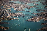 An aerial view of the city of Sydney Australia. In the photo you can see Darling Harbor, the Harbor Bridge, and the Sydney Opera House. There are many boats in the harbor leaving white ripples in the water.
