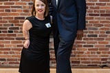 A white redheaded dwarf woman in a black dress standing next to a tall African-American man wearing a blue suit.