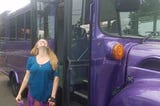 Smiling white woman with long strawberry blonde hair wearing a blue scoop neck shirts looks up smiling in front of a purple sparkly schoolbus.