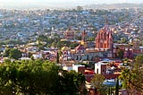 Hilltop view of a city spread out below, with trees in the foreground. There is a large pink stone church with steeples