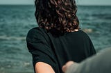 Photo of a woman walking away from the camera, towards to water and blue sky. Her arm is behind her, holding the hand of someone who’s are is reaching forward to her.