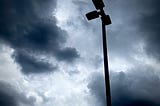 A silhouette of a parking lot streetlight with three bulbs, with a background of dark ominous clouds