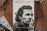The cover of the novel “Green Lights” by Matthew McConaughey situated on a wooden table next to a pair of glasses and a stack of books.