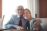 Sitting and smiling elderly man with left arm around seated and smiling younger woman who is a family member