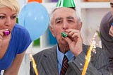 Senior man wearing grey suit celebrating his 70th birthday with green party hat on blowing green party favour, accompanied by mid age blonde lady to his left wearing blue blouse blowing pink party favour, another younger man on his right blowing favour wearing move shirt. Balloons and streamers around…