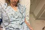 White woman in hospital bed, hospital gown, with an IV in her left arm, and with oxygen in her nose. She looks miserable.