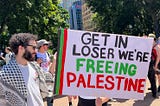 A Palestinian protestor holding a sign stating, “Get In Loser We’re Freeing Palestine”