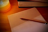 Some sheets of blank paper on a wooden surface. There is a wooden pencil on the papers, a stack of books in the background, and there is a lit candle providing a soft glow in the corner.