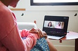 A woman conducts a teletherapy session on a laptop. The screen of her computer shows two people in a Zoom call.