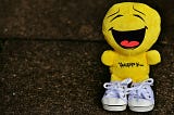 A bright yellow plush toy with a large, joyful laughing face stands on a dark textured surface. The toy has “Happy…” embroidered on its body and is wearing small white lace-up sneakers.
