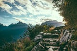 Rocky mountain trail under a partly cloudy sky with shrubbery on either side and peaks in the distance.
