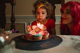 Little girl in a Wonder Woman costume blowing out candles on cupcakes. Mom in a shiny red wig crouched beside her, looking on