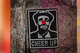 A black and white sticker of sad clown with a red nose and the caption “cheer up”