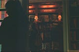 picture of a woman on the left side, looking to the left at some books. In the background, another woman is browsing bookshelves in a dimly lit room.