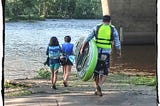 Photo of man carrying a paddle board approaching a river with his two daughters.