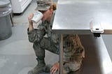 J L Cross enjoying a cup of coffee while she was deployed in Kuwait in year 2017. She’s seen dressed in her AF Airman Battle Uniform sipping from a large white mug. She’s hunkered under a steal kitchen table, an attempt to avoid leadership at the time. Shown in the back ground is the Kitchen in which she worked to serve hundred, if not during some rotations of manning, over a thousand Marines, Navy, Air Force, Army, and other allied international forces at her base.