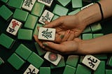 Two hands with a hair tie around one wrist and painted nails hold a single mahjong tile. There are several other tiles scattered on a black surface underneath the pair of hands. Some are face up, and most others are face down, showing a solid green.