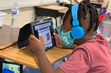 A masked school-aged child wearing headphones looks at a tablet device while sitting at a school desk.