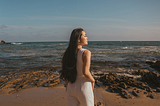 A picture of a Vietnamese American woman standing with her back to the camera, in front of a beach with calm waves. The picture is warm toned. She’s wearing a white jumpsuit with baby blue platform heels, and looks off into the sunset to her right.