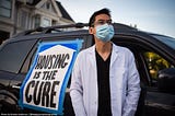 A person in a medical mask and white doctor’s coat stands in front of a vehicle with a sign that reads “HOUSING IS THE CURE”